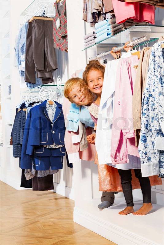 Happy boy and girl play hide-and-seek in clothes together while shopping in the clothes store, stock photo