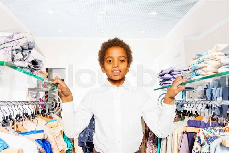 African boy in white shirt stand between hangers with clothes during shopping in the clothes store, stock photo