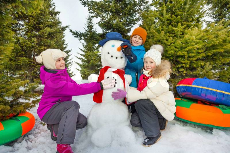 Smiling children make cute snowman with boy holding his carrot nose during beautiful winter day in the forest, stock photo