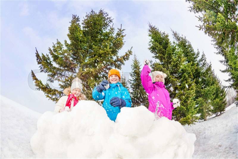 Girls and boy playing snowballs game together standing behind the snow wall with fir forest on the background during winter day, stock photo