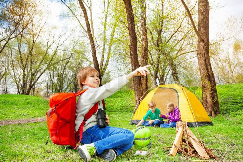 Happy boy with red backpack points with finger near the wooden bonfire and yellow tent and other kids during camping in summer weather, stock photo