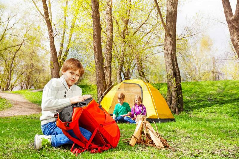 Boy packing the things into red rucksack near the wooden bonfire and his friends sit at yellow tent during camping in summer weather, stock photo