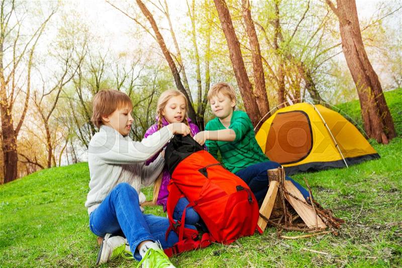 Three kids packing the things into red rucksack near the wooden bonfire and yellow tent during camping in summer weather, stock photo