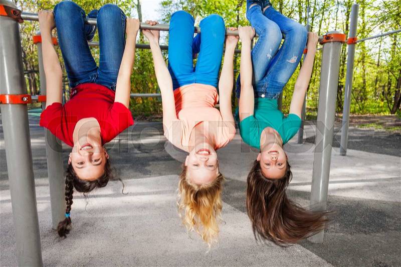 Three girls hanging upside-down on the brachiating bar at the sports ground during summer day with trees on background, stock photo