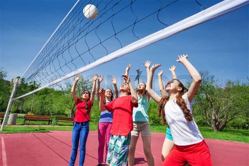 Group of teens with arms up play volleyball near the net on the court during sunny summer day outside, stock photo