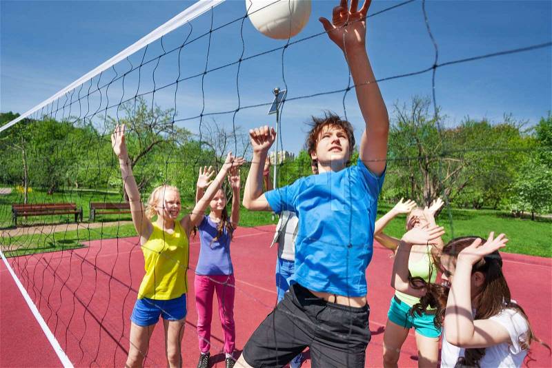 Jumping boy trying to catch ball with arms up plays volleyball with other teens near net on the playing court during sunny summer day, stock photo