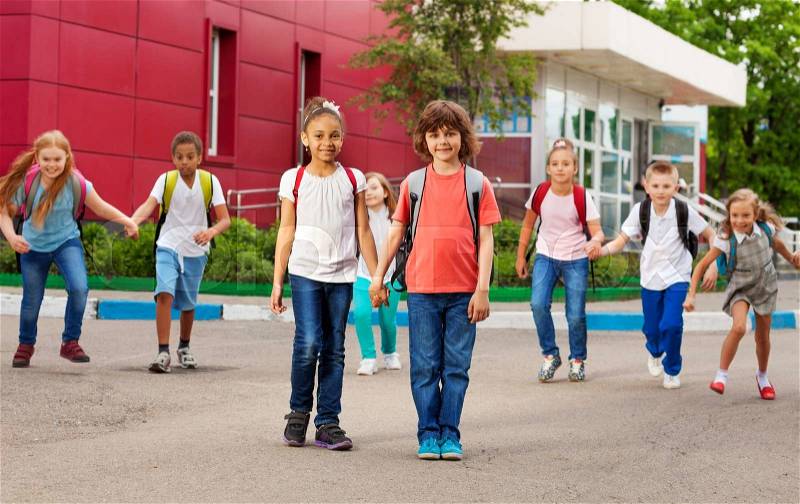 Rows of kids with rucksacks near school walking holding hands during summer day time, stock photo