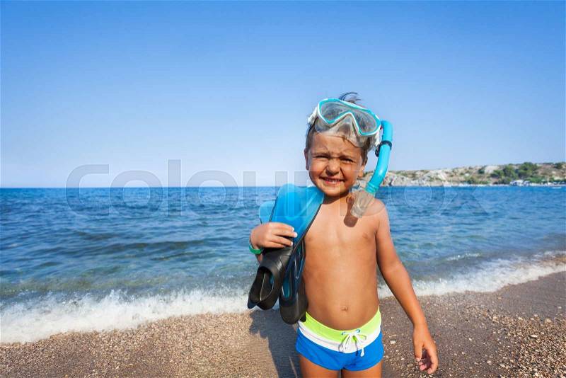 Boy with scuba mask and paddles stands on the seashore squinting against the sun, stock photo