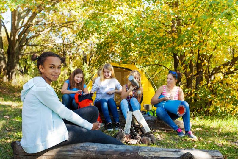 Smiling African girl sitting on log with other girls behind her at camping, stock photo