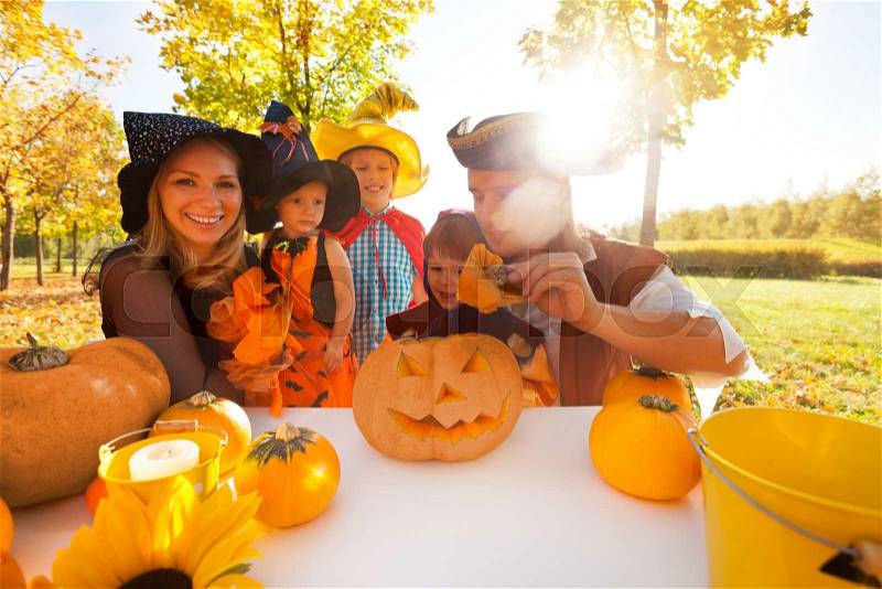 Family in Halloween costumes crafting Jack-O\'-Lantern from pumpkin sitting at the table outside during beautiful sunny autumn day, stock photo