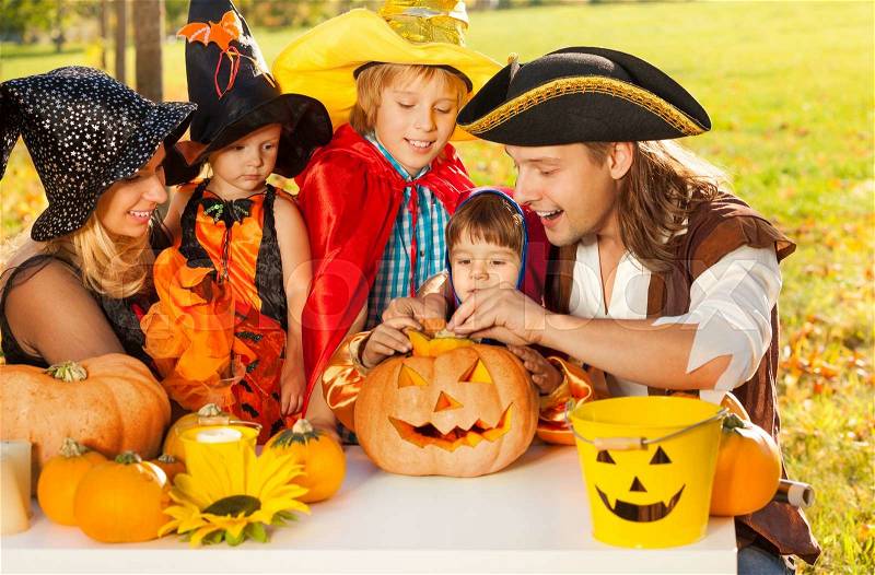 Family crafting Jack-O\'-Lantern from pumpkin in Halloween costumes sitting at the table outside during beautiful sunny autumn day, stock photo