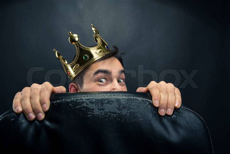 Scared man with crown on the head hidding behind the chair, stock photo
