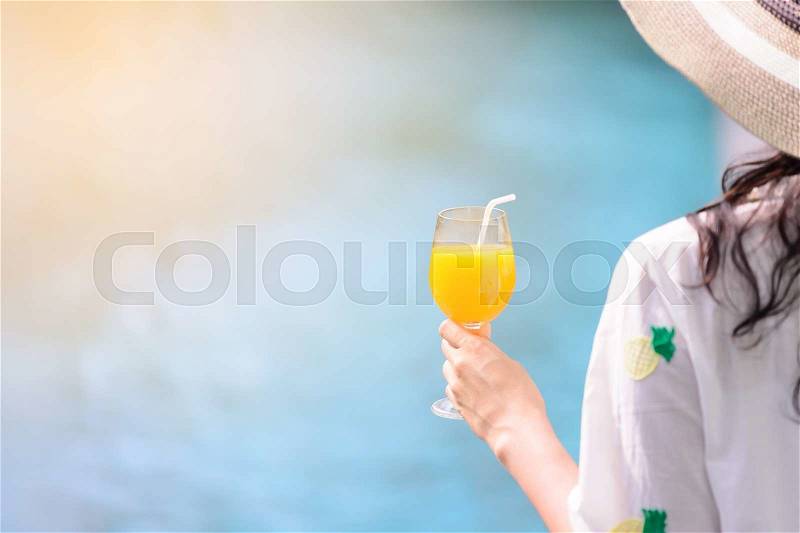 Rear view of beautiful woman in hat is holding orange juice glass at poolside in summer, stock photo