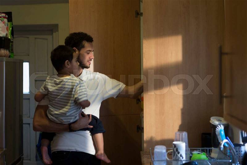 Young father in the kitchen, getting something out of the cupboard with one hand with his son on his shoulder, stock photo