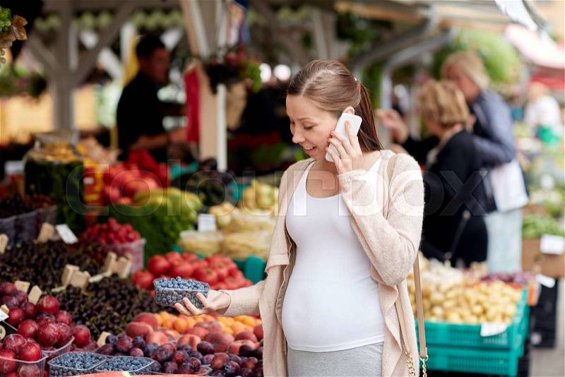 Sale, shopping, food, pregnancy and people concept - happy pregnant woman choosing berries and calling on smartphone at street market, stock photo