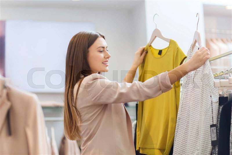 Sale, clothes , shopping, fashion and people concept - happy young woman choosing between two shirts in mall or clothing store, stock photo