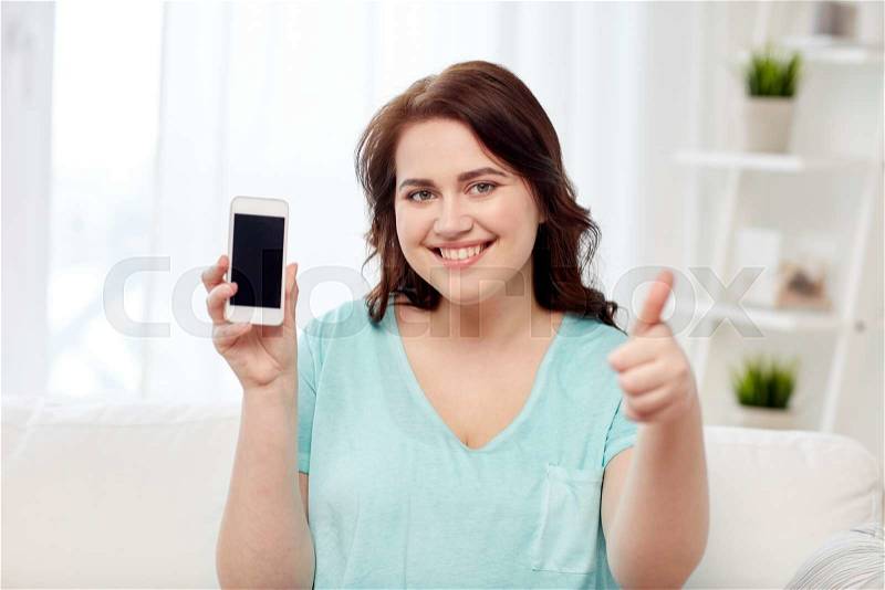 People, technology, communication and leisure concept - happy young plus size woman sitting on sofa and showing smartphone blank screen and showing thumbs up at home, stock photo