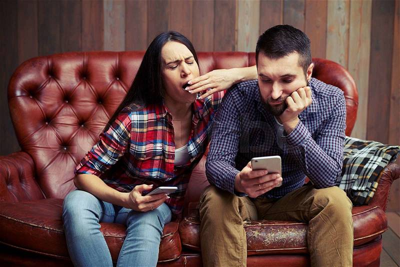 Boring couple sitting on sofa and looking at smartphone, stock photo