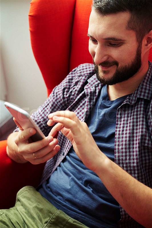 Cheerful young man resting on chair and using his smartphone at home, stock photo
