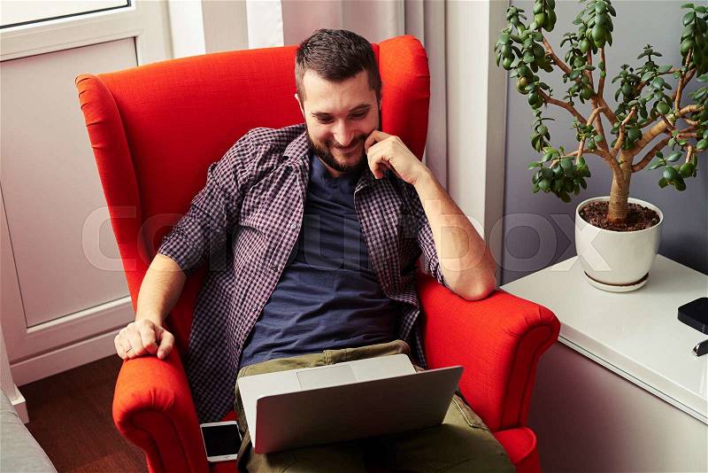 Smiley young man sitting in the chair and looking at laptop, stock photo