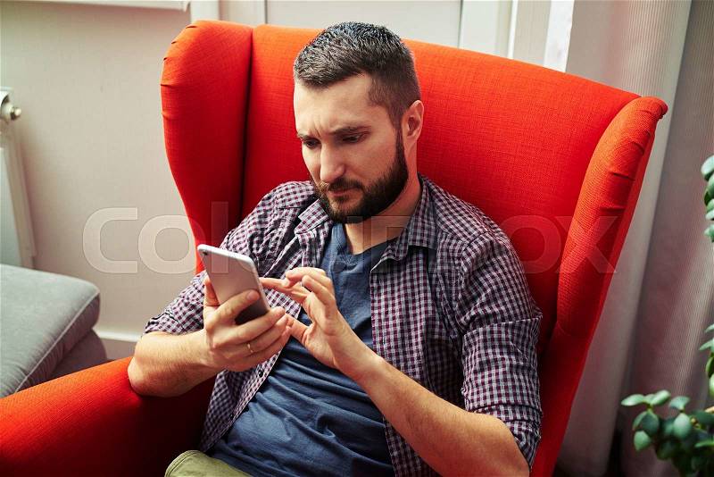 Serious young man sitting on the chair and using his smartphone, stock photo