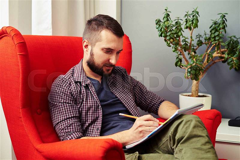 Serious young man sitting on the chair and writing in the notebook, stock photo