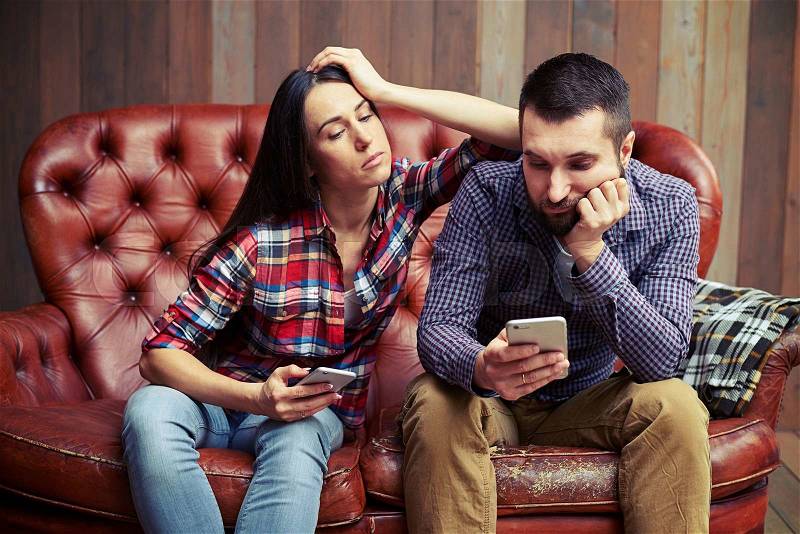 Sad couple sitting on sofa and looking at smartphone , stock photo