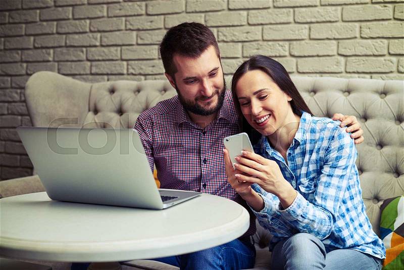 Young smiley couple sitting on couch and looking at smartphone, stock photo