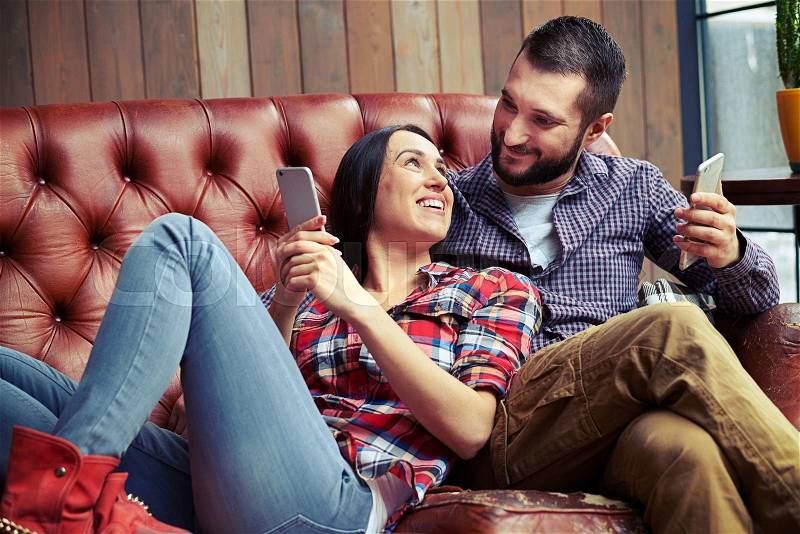 Smiley man and woman resting on sofa and using smartphones, stock photo