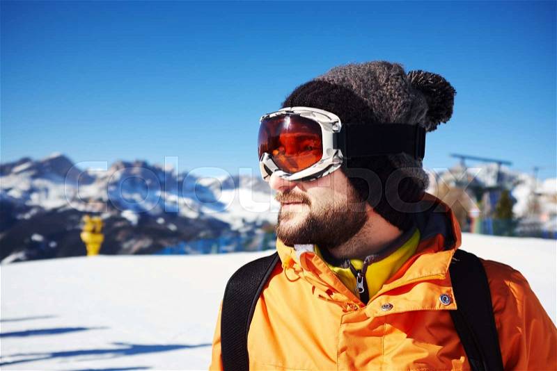 Sideview portrait of smiley skier in orange jacket and mask over mountain view, stock photo