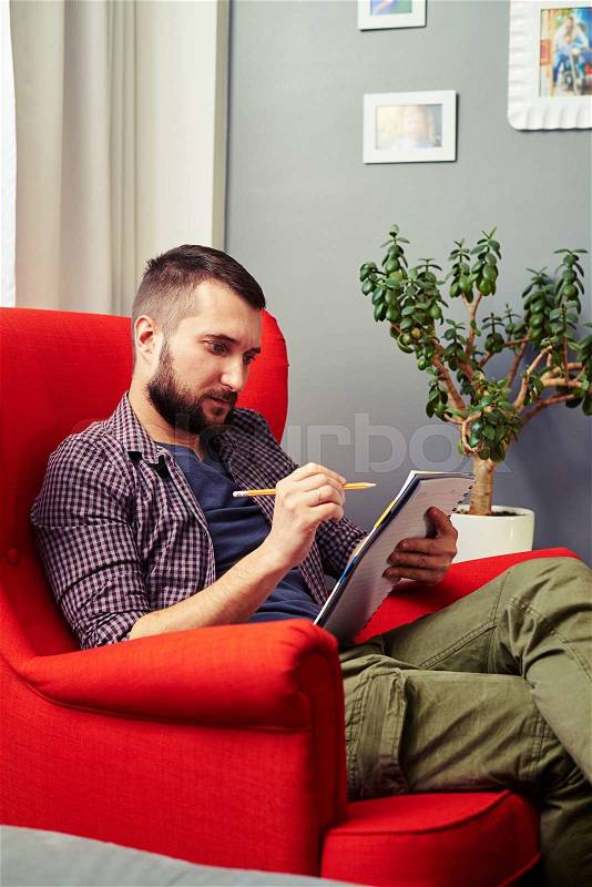 Serious young man with writing pad sitting on the chair at home, stock photo