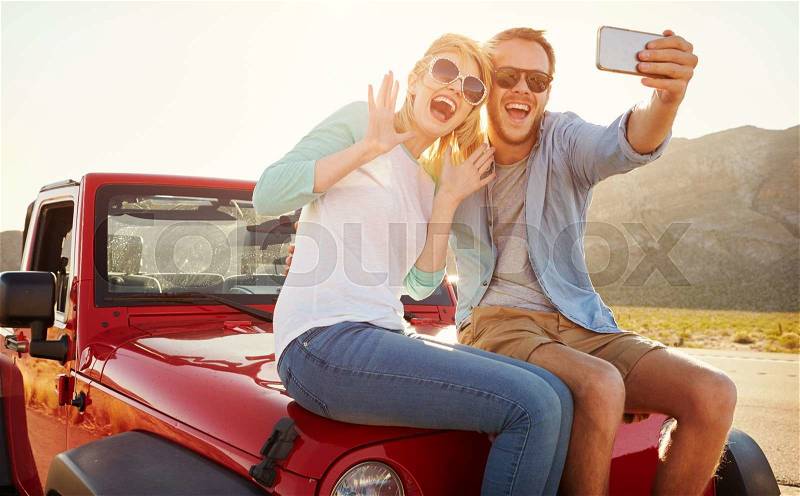 Couple On Road Trip Sit On Convertible Car Taking Selfie, stock photo