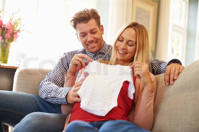 Pregnant Couple On Sofa At Home Looking At Baby Clothes, stock photo