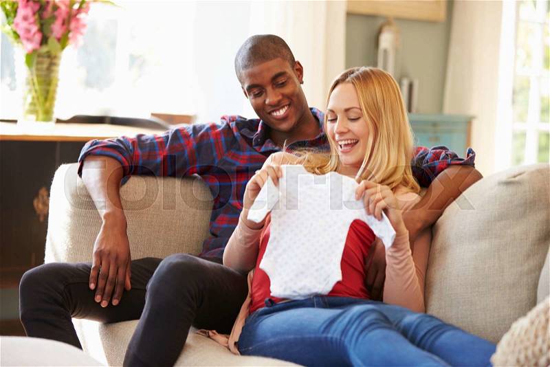 Pregnant Couple On Sofa At Home Looking At Baby Clothes, stock photo