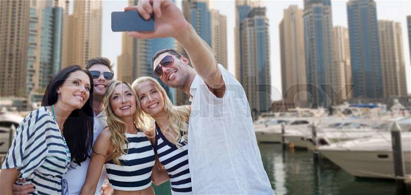 Summer, sea, tourism, technology and people concept - group of smiling friends with smartphone taking selfie over dubai city harbor or waterfront background, stock photo