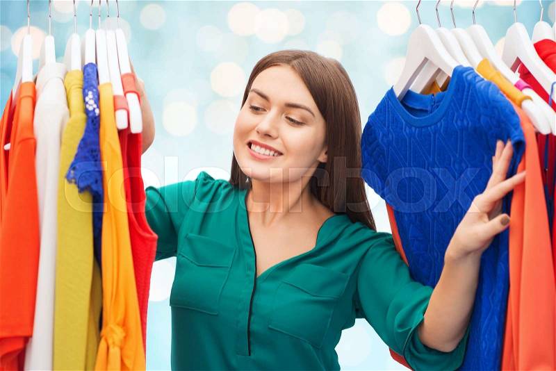 Clothing, fashion, style and people concept - happy woman choosing clothes at wardrobe over blue holidays lights background, stock photo