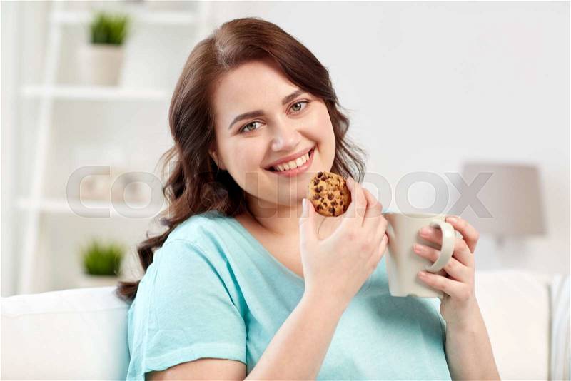 People, junk food, unhealthy eating and leisure concept - happy plus size young woman with cup of tea with cookie at home, stock photo