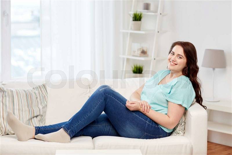 People and leisure concept - happy young woman plus size sitting on sofa at home, stock photo