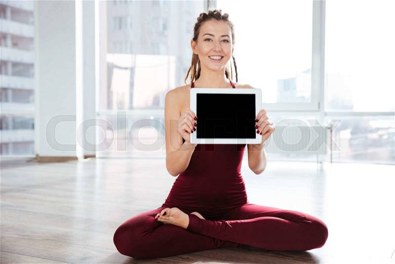 Happy attrative young woman with dreadlocks sitting in yoga pose and showing blank screen tablet, stock photo