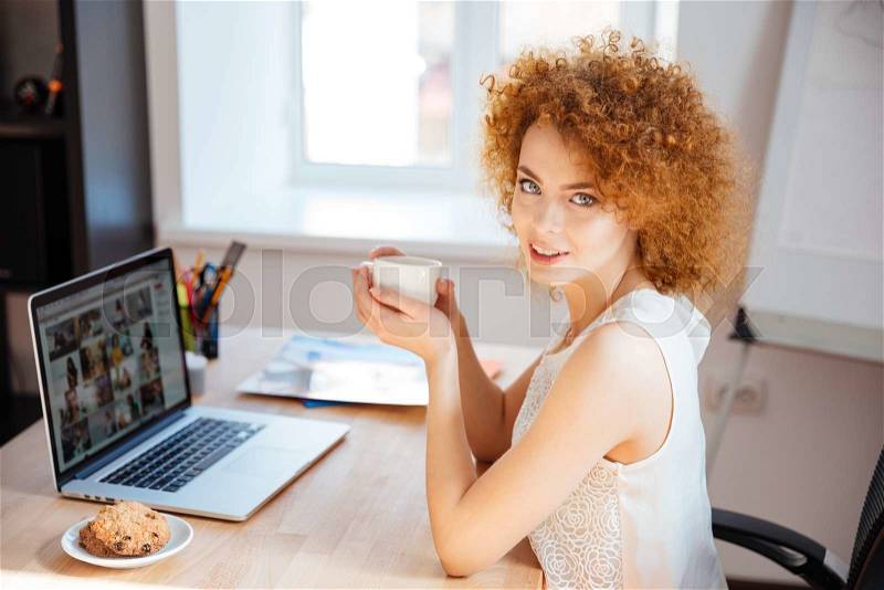 Young woman drinking coffee at the table and using laptop in the oofice, stock photo
