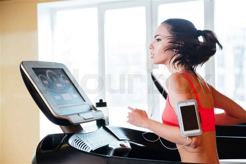 Attractive concentrated young woman running on treadmill in gym and listening to music from blank screen mobile phone , stock photo