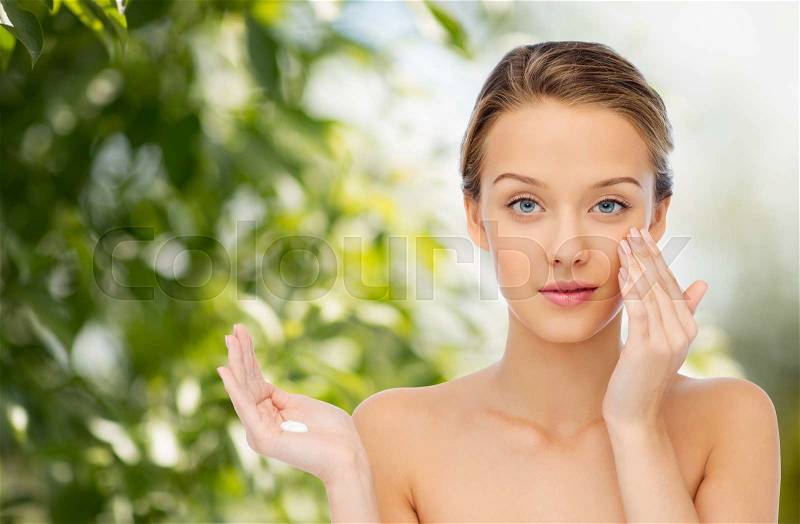 Beauty, people, cosmetics, skincare and health concept - young woman applying cream to her face over green natural background, stock photo