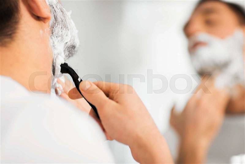 Beauty, hygiene, shaving, grooming and people concept - close up of young man looking to mirror and shaving beard with manual razor blade at home bathroom, stock photo