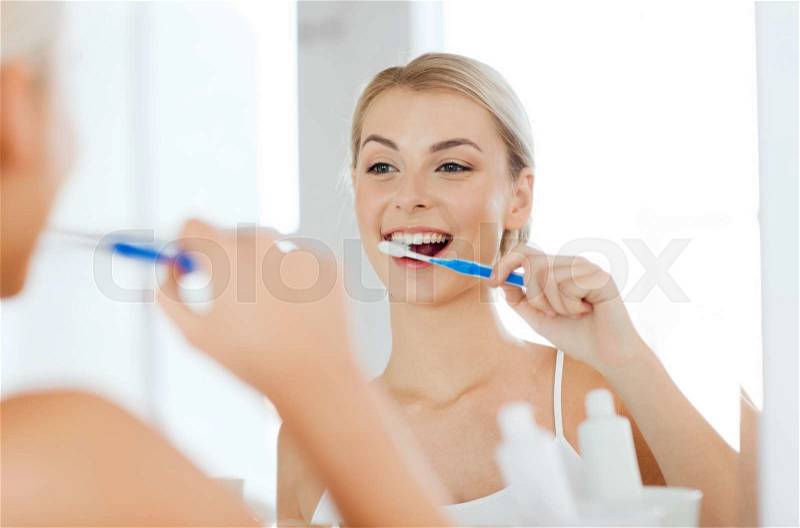Health care, dental hygiene, people and beauty concept - smiling young woman with toothbrush cleaning teeth and looking to mirror at home bathroom, stock photo