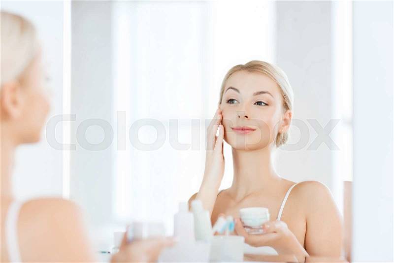 Beauty, skin care and people concept - smiling young woman applying cream to face and looking to mirror at home bathroom, stock photo