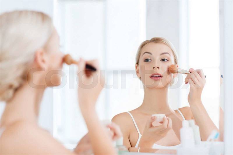 Beauty, make up, cosmetics, morning and people concept - smiling young woman makeup brush and powder foundation looking to mirror at home bathroom, stock photo