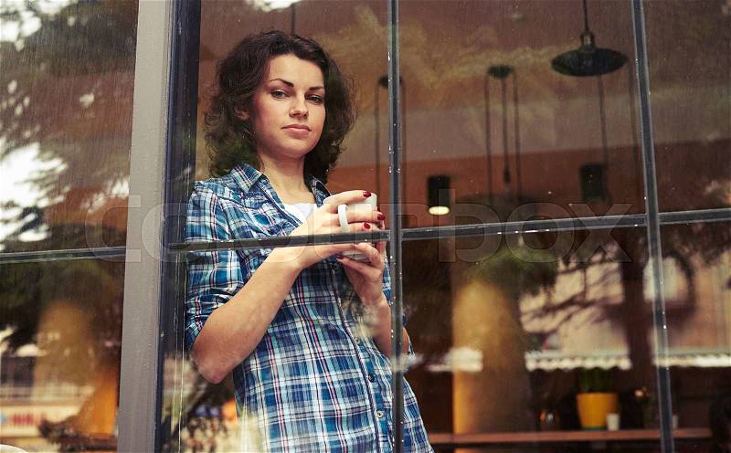 Beautiful girl standing near window, holding cup of coffee and looking at camera, stock photo