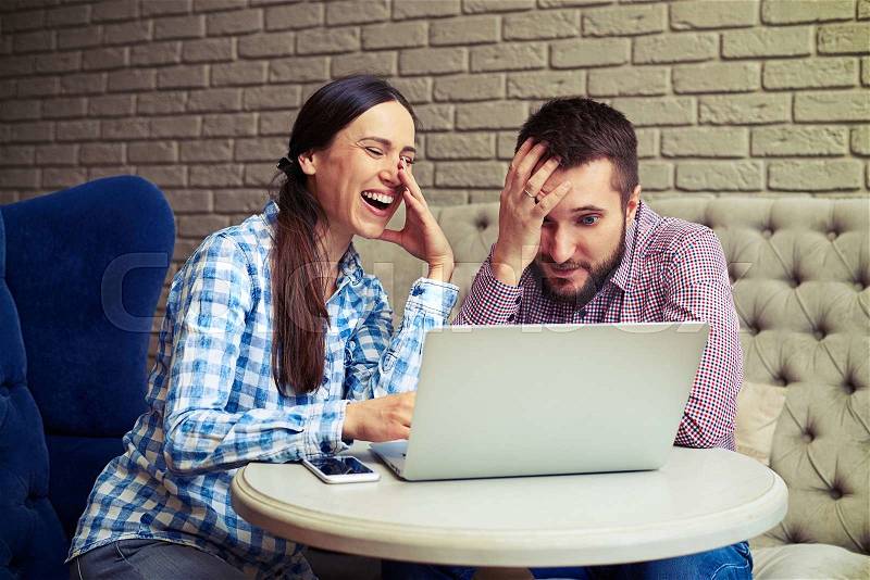 Laughing woman and sad man sitting on sofa and looking at laptop, stock photo