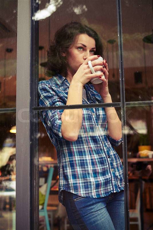 Beautiful young woman standing near big window and drinking the coffee, stock photo