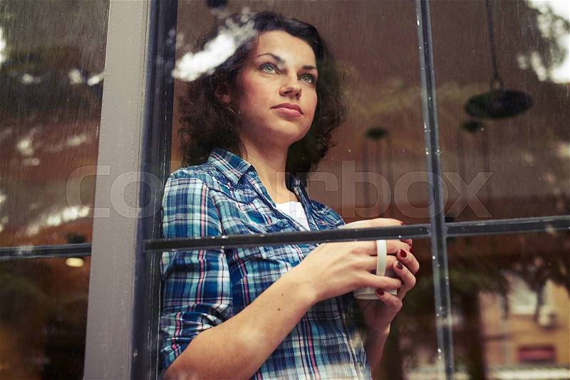 Beautiful young woman standing near the window, holding cup of coffee and looking into the distance, stock photo